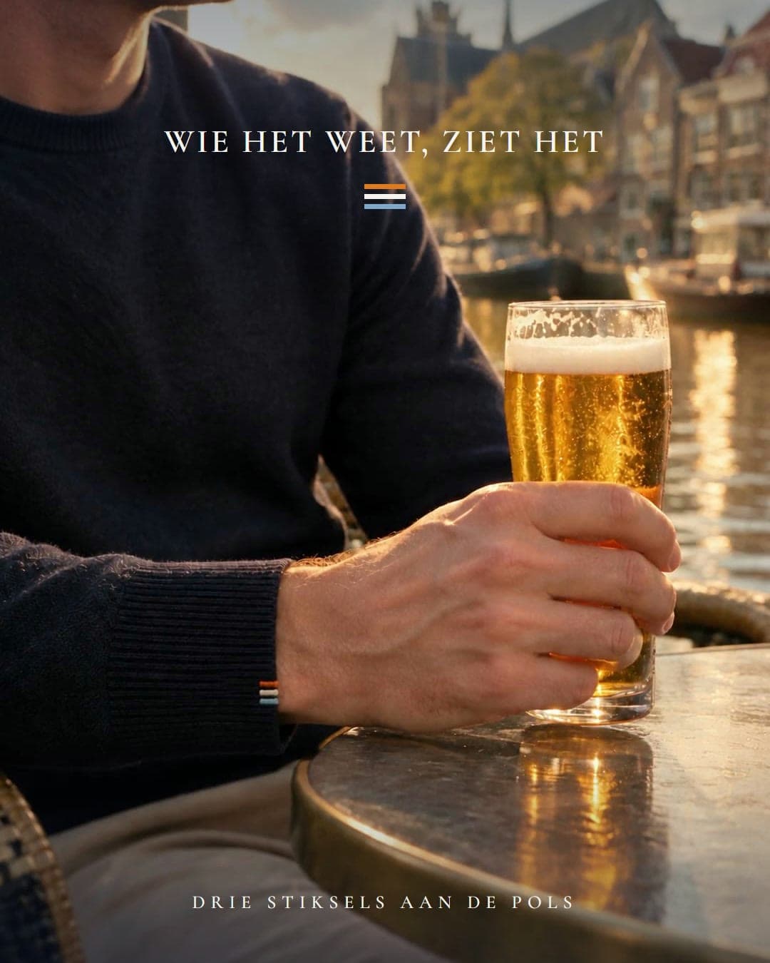 Een man in een navy trui houdt een glas bier vast op een terras aan het water; aan zijn manchet drie stiksels in oranje, wit en blauw.